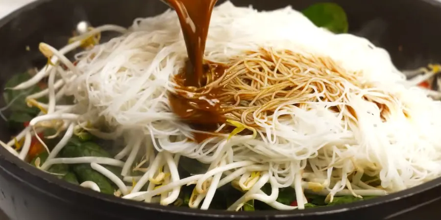 Dark savory stir-fry sauce being poured over rice vermicelli and bean sprouts in a wok