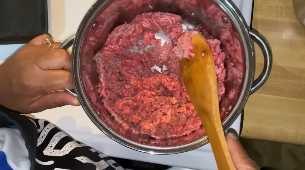 Raw ground beef being broken up with a wooden spoon in a stainless steel pot