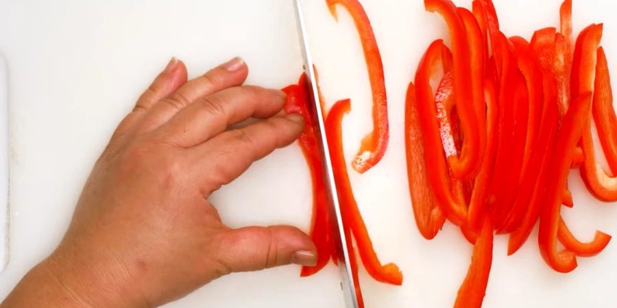 Knife cutting red bell pepper into thin strips on a white cutting board