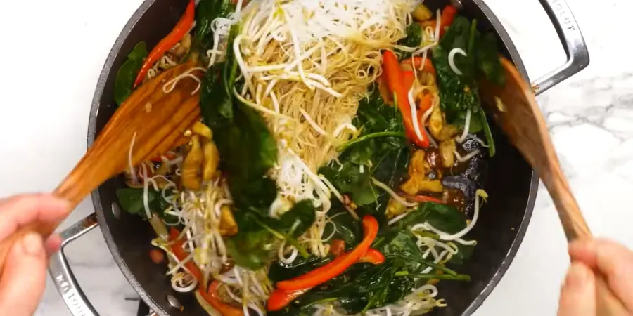 Tossing thin rice vermicelli with red bell pepper and bean sprouts in a wok using two wooden spatulas