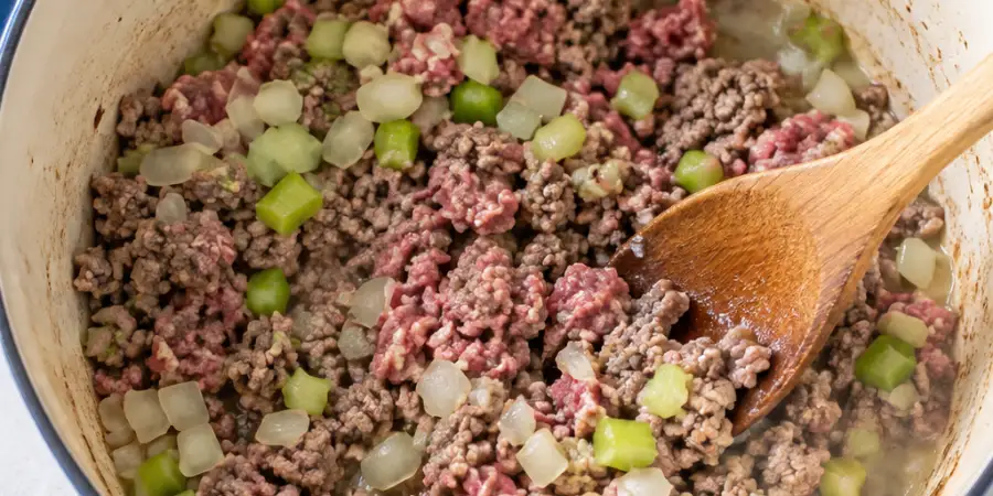 Ground beef, onion, and celery browning in the pot, being broken up with a wooden spoon