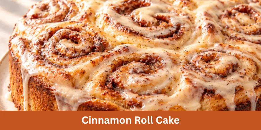 A whole homemade cinnamon roll cake is presented on a simple serving surface in a warm home kitchen. The finished cake is shown from a 45-degree angle with its baked cinnamon-swirl surface and complete shape clearly visible. The softly blurred kitchen background creates a warm and natural recipe setting.