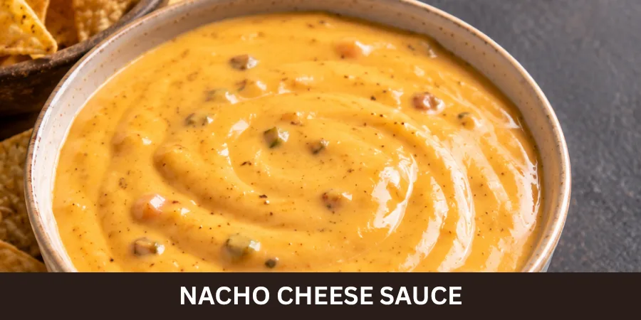 A bowl of smooth homemade Nacho Cheese Sauce sits beside a small serving of tortilla chips on a dark neutral surface, shown from a 45-degree angle with soft lighting.