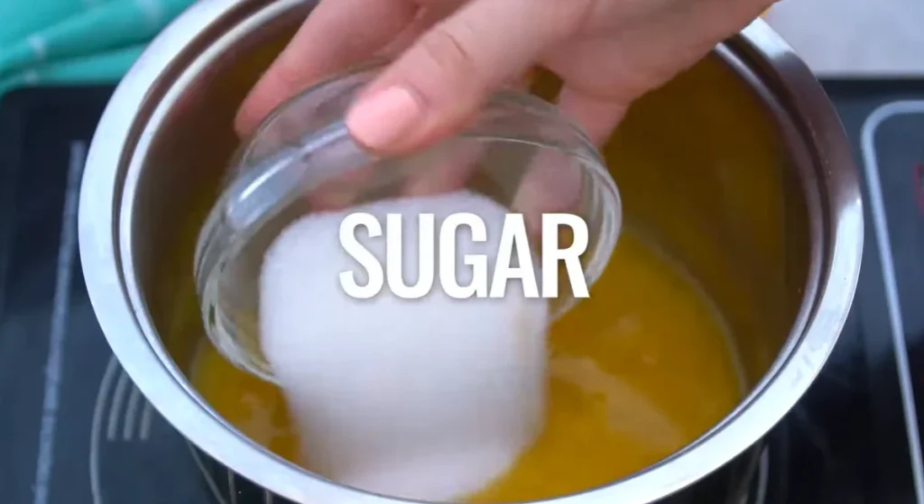 Adding granulated sugar from a small glass bowl to orange juice in a saucepan