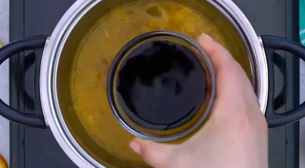 Adding low-sodium soy sauce from a small glass bowl into the saucepan for orange sauce