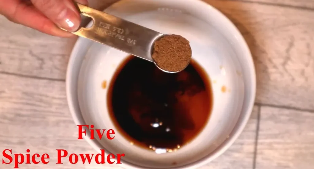 Half-teaspoon measuring spoon filled with Chinese five-spice powder over a bowl of dark sauce ingredients