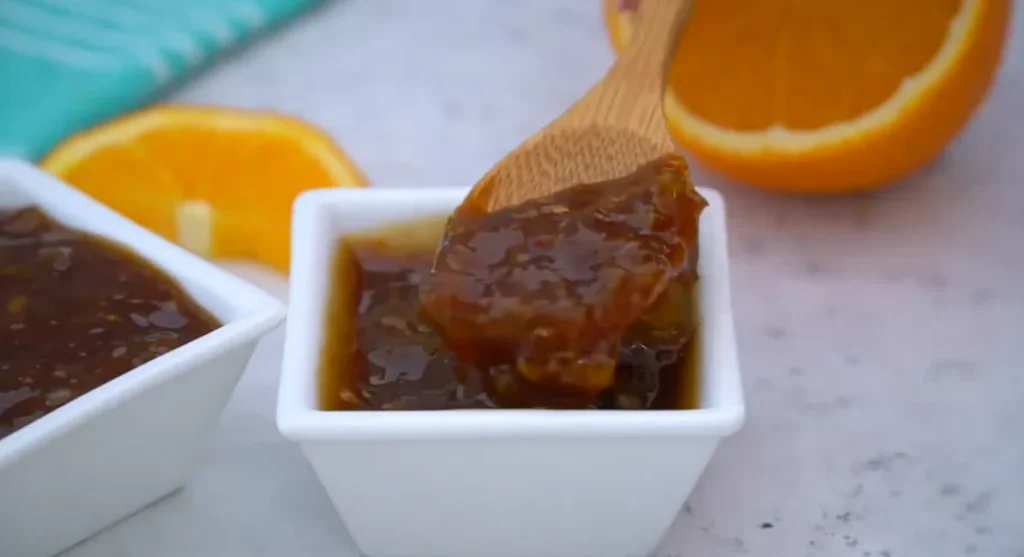 Homemade orange sauce in a white square bowl with a wooden spoon lifting thick, glossy sauce