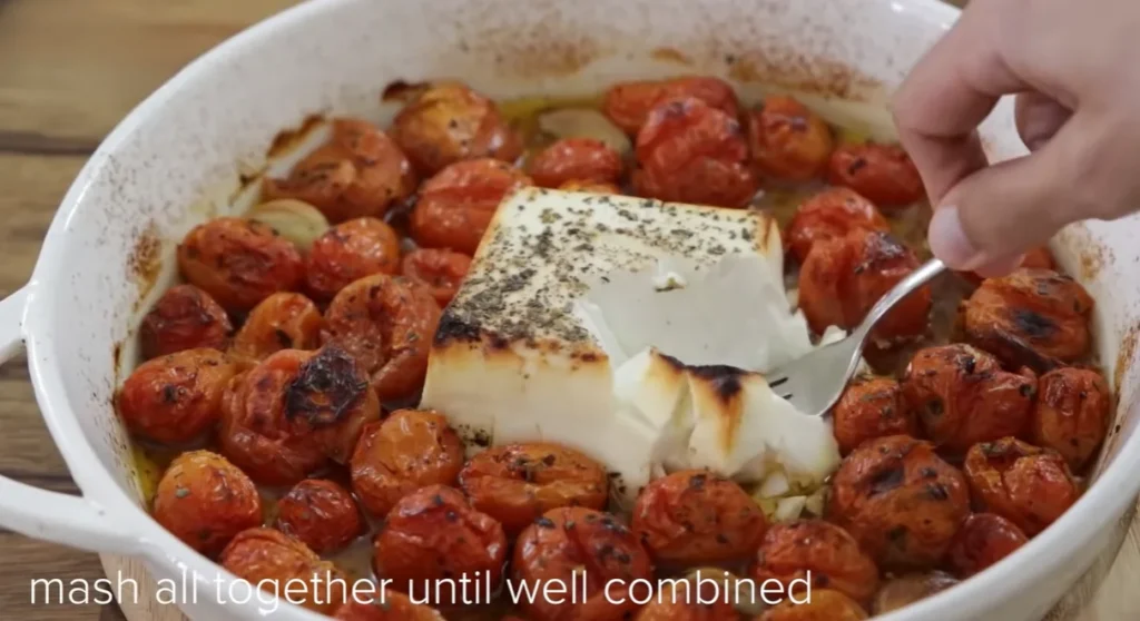 Mashing roasted feta with burst cherry tomatoes and garlic using a fork