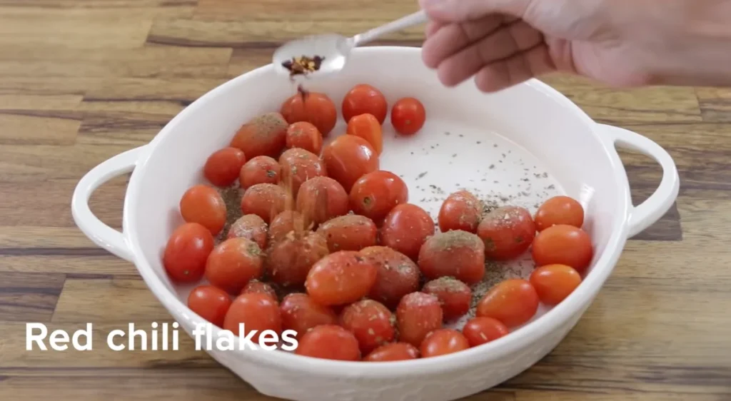 Red chili flakes being added to seasoned cherry tomatoes with a spoon